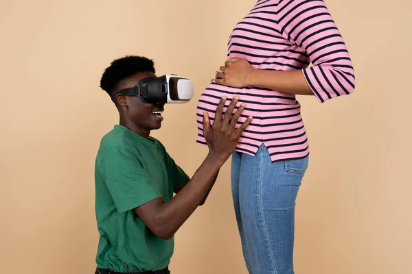 Glad surprised young black father in vr glasses hold woman belly, isolated on beige background, studio, close up, cropped. Parents care, family, relationships and pregnancy, fun and entertainment