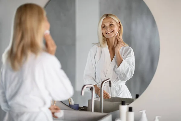 バスルームでコットンパッドの近くの鏡で肌をクレンジングする美しい成熟した女性の肖像画 彼女の反射に微笑む魅力的なシニアウーマン 自宅でセルフケアルーチンを楽しむ 選択的な焦点 — ストック写真