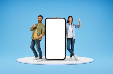 Mobil uygulama. Olumlu, çoklu etnik çeşitlilikte arkadaşlar beyaz ekranlı büyük bir telefonla poz veren Asyalı genç adam ve beyaz bir kadın, başparmak kaldırıyor, gülümsüyor, mavi arka planda izole edilmiş, boş bir alan.