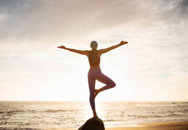 Spor giyimli genç beyaz kadın taş üzerinde yoga yapıyor, nefes egzersizlerinin, deniz sahilinde dengenin ve özgürlüğün tadını çıkarıyor. Sabah antrenmanı, spor, vücut bakımı ve Asana.