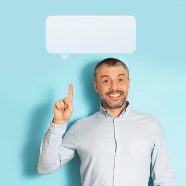 Heyecanlı orta yaşlı beyaz iş adamı işaret parmağını boş beyaz iletişim balonuna doğrultuyor ve mavi stüdyo geçmişine gülümsüyor. Güzel bir iş fikri veya tavsiyesi var.