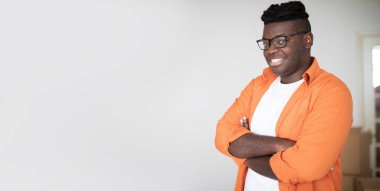 Gülümseyen Afrikalı genç Amerikalı adam, kollarını katlayıp karton kutulara yaslanmış şekilde poz veren, yakışıklı siyahi adam, taşınma gününde New Home 'da duruyor, panorama ve fotokopi alanı.