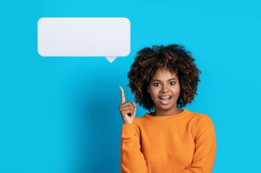 Harika fikir, ilham konsepti. Heyecanlı siyahi kadının portresi parmağını kaldırıyor, boş konuşma balonu gösteriyor ve gülümsüyor, yaratıcı bir fikri var, mavi stüdyo arka planında duruyor.