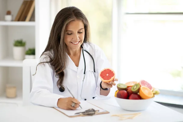 Glad european middle aged woman doctor nutritionist makes notes of diet plan at table with fruits, holds grapefruit in clinic office interior. Work, professional weight loss and health care