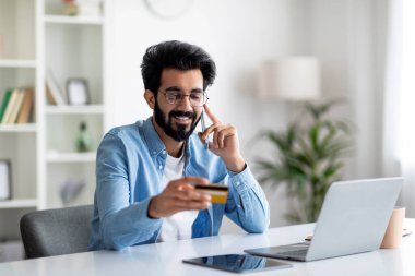 Gülümseyen Hintli genç adam seyahat biletlerini internetten alıyor, cep telefonuyla konuşuyor ve kredi kartı tutuyor, İç İşleri 'nde otururken, mutlu doğu adamı cep telefonu bankacılığının keyfini çıkarıyor, kopyalama alanı kopyalıyor.