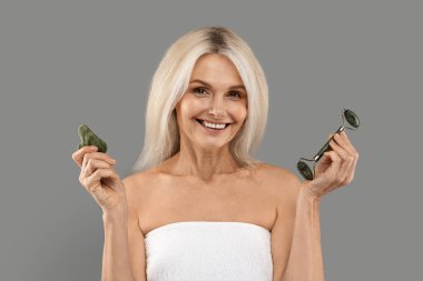 Cilt Aletleri. Gri arka planda dikilirken kameraya gülümseyen, cilt bakımını yapan, guasha taşı kazıyıcısı ve yeşim yüzlü çekici bir kadın.