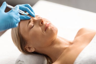 Kozmetik doktoru güzellik salonunda olgun bir kadına yaşlanma karşıtı iğneler yapıyor. Orta yaşlı çekici bir kadın kırışıklıklara karşı tedavi görüyor. Kaşlar arası bölgede güzellik aşısı oluyor.