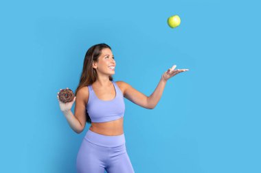 Elinde donutla gülümseyen beyaz kadın yeşil elma kusuyor, eğleniyor, mavi stüdyo arka planında soyutlanıyor. Diyet, şeker ya da meyve seçimi, vücut bakımı, kilo kaybı