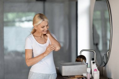 Vücut Bakımı Konsepti. Gülümseyen Olgun Kadın Nemlendirici Losyon Sürerken Evde Modern Banyo 'da Beklerken, Çekici Mutlu Son Sınıftaki Kadın Besleyici Krem Uyduruyor, Boşluğu Kopyala