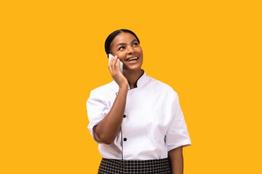 Üniformalı neşeli siyah aşçı cep telefonuyla konuşuyor, gülümseyen Afrikalı Amerikalı kadın aşçı, sarı stüdyonun arka planında yalnızken keyifli bir telefon görüşmesi yapıyor, boşluğu kopyalıyor.
