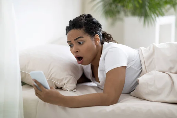 Late For Work. Shocked Overslept Black Woman Looking At Smartphone In Her Hand, Worried African American Lady Lying In Bed, Having No Time To Get Ready In The Morning, Suffering Tardiness, Closeup