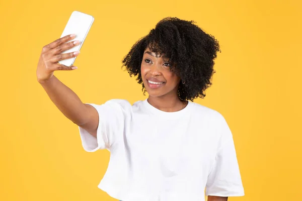 stock image Cheerful young black curly lady in white t-shirt makes selfie on smartphone for social media, blog, isolated on orange background, studio. Ad and offer, video call, blogger