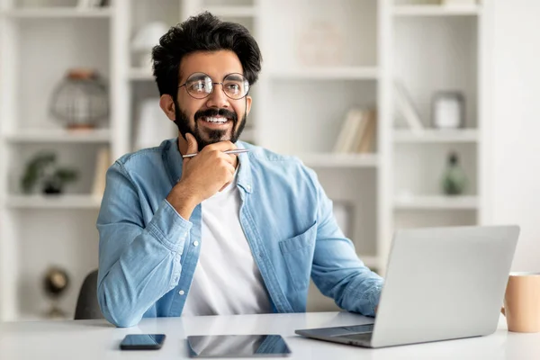 Dreamy Young Indian Man Sitting At Desk With Laptop In Home Office And Looking Away, Smiling Eastern Male Freelancer Thinking About Business Plans Or Having Inspiration Moments, Touching Chin