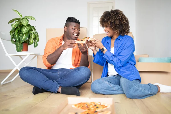Black Millennial Couple With Pizza Celebrating Moving To New Home, Cheerful Young African American Spouses Sitting On Floor Among Cardboard Boxes, Enjoying Lunch Break And Having Fun Together