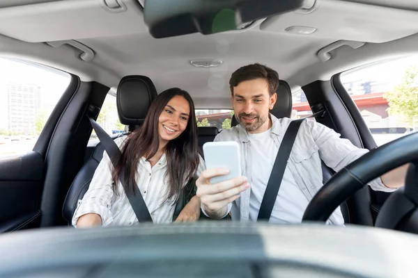Happy millennial man and woman having car trip together, using cell phone and smiling, copy space. Cheerful couple travellers enjoying journey by cozy auto, tracking their way on smartphone