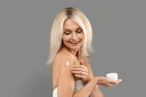 美丽的成熟金发女人在灰蒙蒙的背景下将润肤霜应用于肩部 具有诱惑力的高级女装保鲜乳罐 享受抗衰老护理 复制空间 — 图库照片