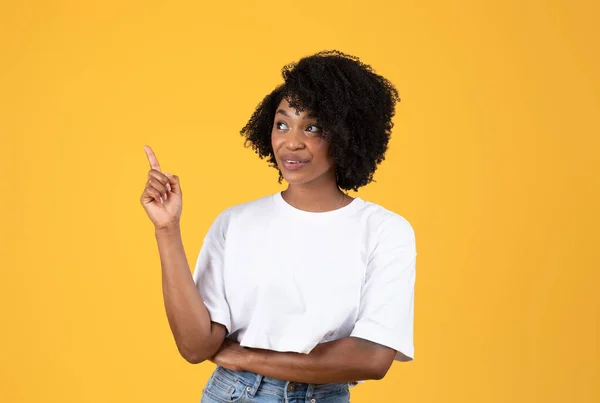 穿着白色T恤的快乐而沉思的年轻黑人卷曲女士在想 手指指向空旷的空间 被橙色背景隔离 工作室 人类的情感 广告和提供 — 图库照片