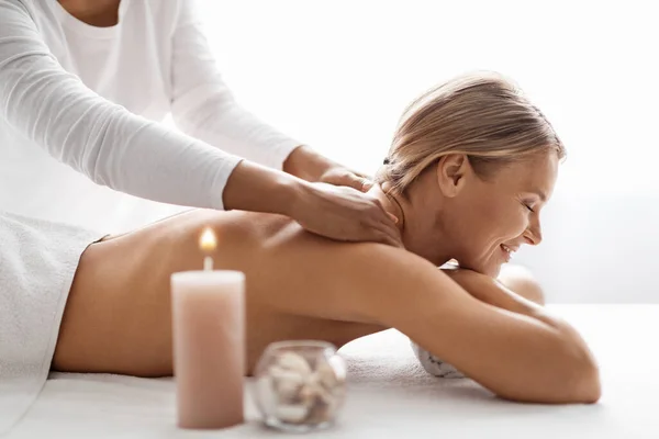 Beautiful middle aged woman having relaxing full body massage at spa salon, female therapist rubbing shoulders of smiling lady, doing wellness beauty treatment to relaxed woman lying on table