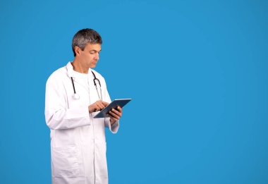 Beyaz önlüklü doktor stüdyoda mavi arka planda poz vermek için dijital tablet bilgisayar kullanıyor. Tıbbi reklam için fotokopi alanının yanında duran gri saçlı pratisyen.