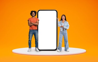 Neşeli, çok kültürlü genç bir çift beyaz ekranlı büyük bir telefonla poz veriyor, güzel mobil uygulama veya harika bir internet teklifi sunuyor, fotokopi alanı, turuncu arka plan.