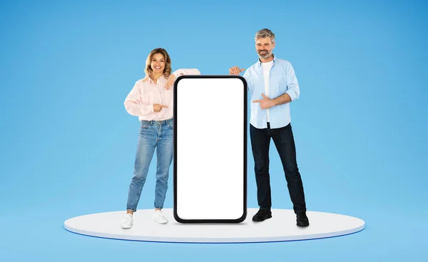 两人花了不少钱快乐的中年男女夫妻站在圆形平台上 手持大手机 屏幕为白色 工作室背景为蓝色 手机应用的空白空间为模特儿 — 图库照片