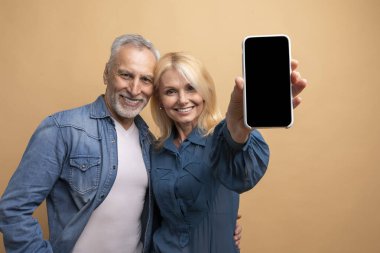 Harika bir mobil uygulama. Neşeli, sevgi dolu, yaşlı çift ve karı koca gündelik kıyafetler giyip siyah ekranlı büyük bir telefon, model, bej renkli stüdyo arka planında izole edilmiş, boş bir yer gösteriyorlar.