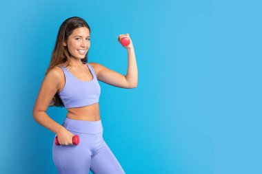 Bin yıllık Avrupalı spor elbiseli bir bayan, dambıllarla kol egzersizi yapıyor, pazılarını gösteriyor, mavi stüdyo arka planında izole edilmiş. Form, motivasyon, spor ve egzersiz, reklam ve teklif