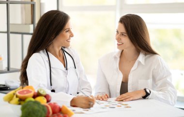 Beyaz önlüklü, mutlu, beyaz tenli beyaz doktor beslenme uzmanı ofis içinde organik sebzeleri ve meyveleri olan genç bir bayana tavsiyede bulunuyor. Sağlık, muayene, diyet planı, profesyonel danışma
