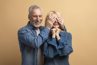 Neşeli, yakışıklı, son sınıf öğrencisi, karısı için doğum günü sürprizi hazırlıyor, yaşlı koca, gülümseyen heyecanlı kadın gözlerini kapatıyor, yıldönümünü birlikte kutluyorlar, bej arka plan.