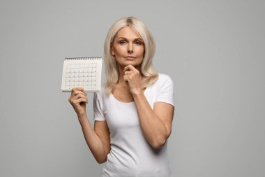 Pensive Mature Lady Holding Blank Menstrual Calendar And Touching Chin, Thoughtful Senior Female Suffering Lack Of Menstruation, Having Climacteric Symptoms Or Menopause, Standing On Grey Background clipart