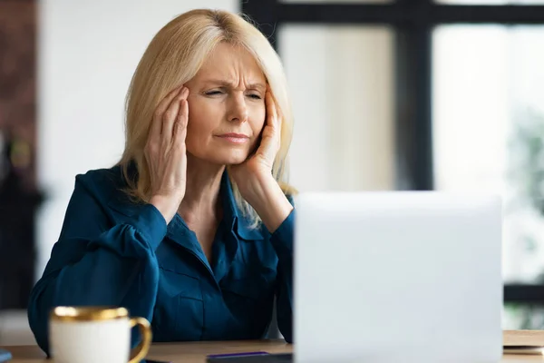 stock image Stressed mature businesswoman having headache while working on laptop, woman looking at screen and rubbing temples, suffering from migraine at home, free space