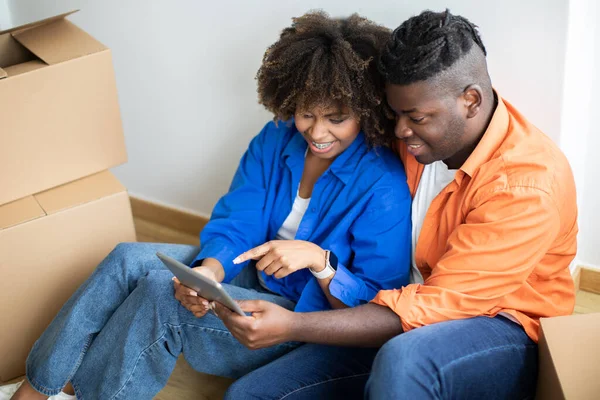 Smiling Black Spouses Moving Home And Using Digital Tablet Together, Happy African American Man And Woman With Modern Gadget Purchasing Furniture Or Planning Design In New Flat, Sitting Among Boxes