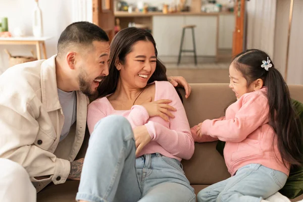 Bonding Moment. Asian family laughs and giggles together as daughter tickles parents for fun, sitting on sofa at home. Kid, mommy and daddy sharing laughter and joy in living room