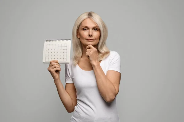 沉思的成熟女士持有空白月历和触动下巴 沉思的老年女性缺少月经 有更年期症状或更年期 站在灰色背景上 — 图库照片