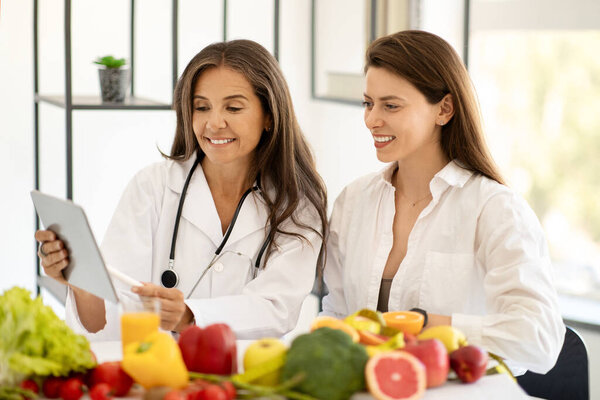 Happy mature caucasian doctor nutritionist in white coat consultation young woman, show plan diet on tablet at table with organic vegetables and fruits in office interior. Health care, weight loss