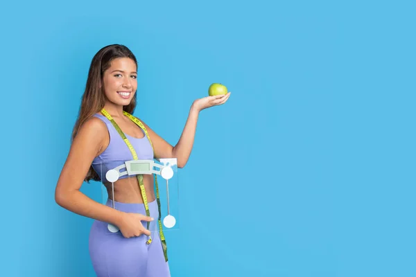 Positive millennial european lady in sportswear with measuring tape hold scales, show green apple, isolated on blue studio background. Fitness, sports, weight loss and diet result, ad and offer