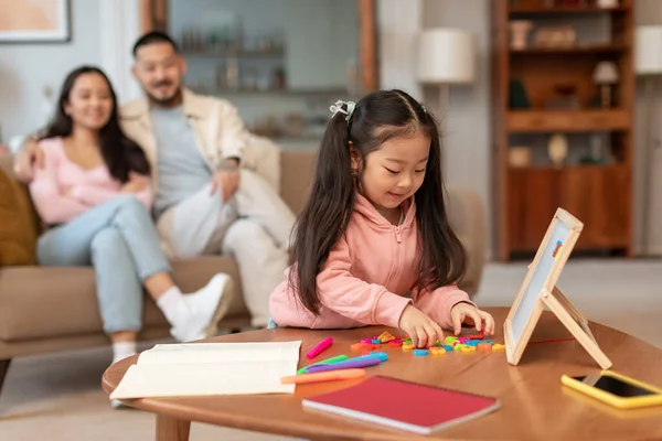 Early Learning Exploration. Little Korean Girl Playing Educational Games, Spelling Words With Alphabet Board At Home, While Parents Relaxing On Sofa In Living Room, Selective Focus