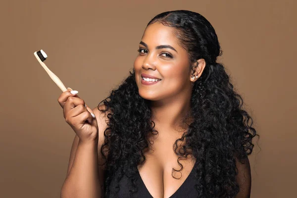 Smiling latin plus size lady holding toothbrush with toothpaste and looking at camera, brazilian woman ready to brush her teeth, standing over brown studio background