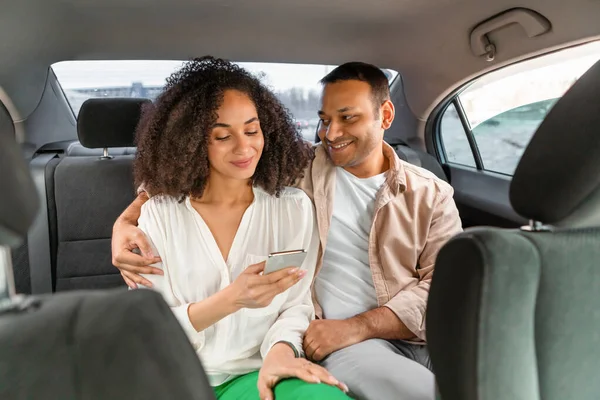 Car Service App. Smiling Middle Eastern Couple in Taxi Embracing And Using Smartphone Promoting Mobile Application for Convenient Transportation, Enjoying Ride Through City Traffic In Auto Inside