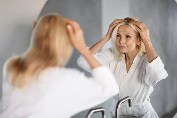 Grey Hair Concept. Upset Mature Woman Looking In Mirror At Her Hair Roots, Worried Senior Lady In Bathrobe Suffering Graying Or Hairloss Problem, Selective Focus On Reflection, Closeup Shot