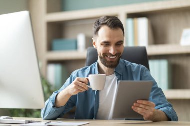 Neşeli genç işadamı çalışma masasında oturuyor, kahve içiyor ve ofiste dijital tablet kullanıyor, yakışıklı girişimci gülümsüyor iş başvurularını kullanıyor ya da haberleri okuyor, işte mola veriyor.