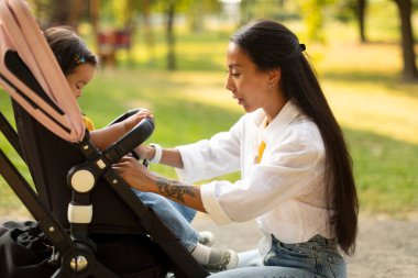 Küçük kızının rahatı için bebek arabasındaki emniyet kemerlerini bağlayan mutlu Koreli anne dışarıda parkta geziyor, yan görüş açısında. Yürüyen Bebek, Annelik ve Bebek Bakımı Konsepti