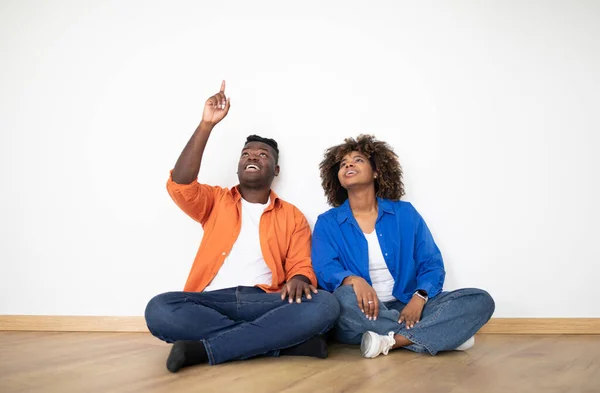 Design Ideas. Excited African American Couple Pointing Up While Sitting On Floor Near White Wall, Smiling Black Spouses Planning Decorations And Furniture In New Home, Celebrating Moving, Copy Space
