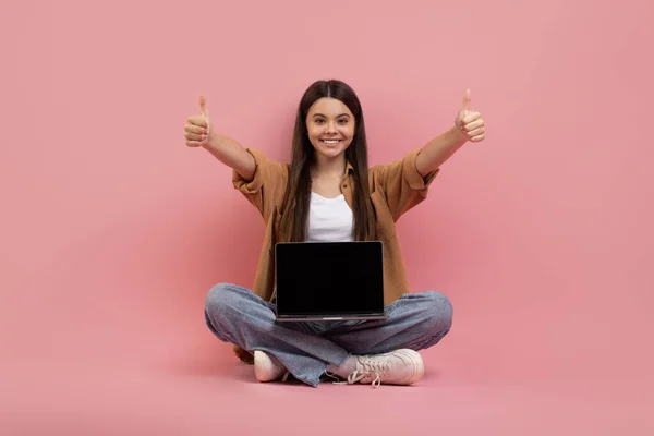 坐在粉红工作室的背景上 微笑着拿着笔记本电脑的少女 带着空白屏幕 大拇指翘起 可爱的少女推荐新网站 模仿设计 — 图库照片