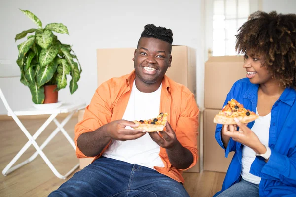 Happy Black Man And Woman Eating Pizza And Celebrating Moving Day, Cheerful Young African American Couple Having Lunch Break Together, Sitting Among Cardboard Boxes In Their New Apartment, Closeup