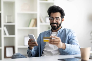 Çevrimiçi Kredi. Akıllı telefon ve kredi kartı kullanan gülümseyen Hintli adamın portresi, Mobil bankacılık için gözlüklü genç ve mutlu bir erkek gözlük takıyor.