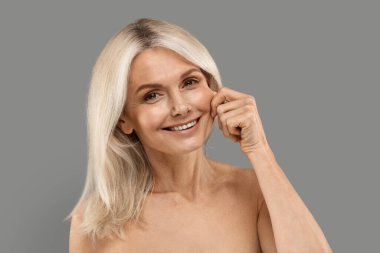 Elmacık kemiği büyütme. Yanağa dokunan ve kameraya gülümseyen güzel olgun bir kadın, gri arka planda ayakta duran estetik cerrahi veya estetik tedavinin mutlu yaşlı kadın örneği.