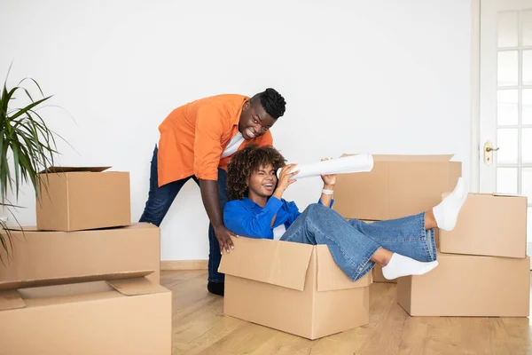 Cheerful Black Couple Having Fun Together While Relocating Home, Positive Young African American Spouses Fooling On Moving Day, Joyful Man Pushing Cardboard Box With His Happy Wife, Copy Space