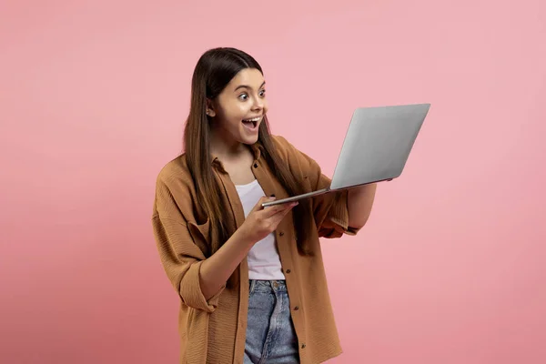兴奋的少女看着笔记本电脑屏幕 吃惊地张开嘴 对大减价或好消息的惊讶少女的肖像 站在粉色工作室的背景上 — 图库照片
