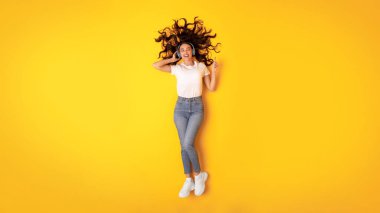 Dijital Müzik Eğlencesi. Heyecanlı genç bayan, kablosuz kulaklıklı müzik dinliyor, parmağıyla işaret ediyor ve sarı stüdyonun arka planında uzanan favori şarkının tadını çıkarıyor.